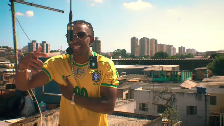 Tchony cantando no microfone na laje, de camisa amarela, com a cidade atrás